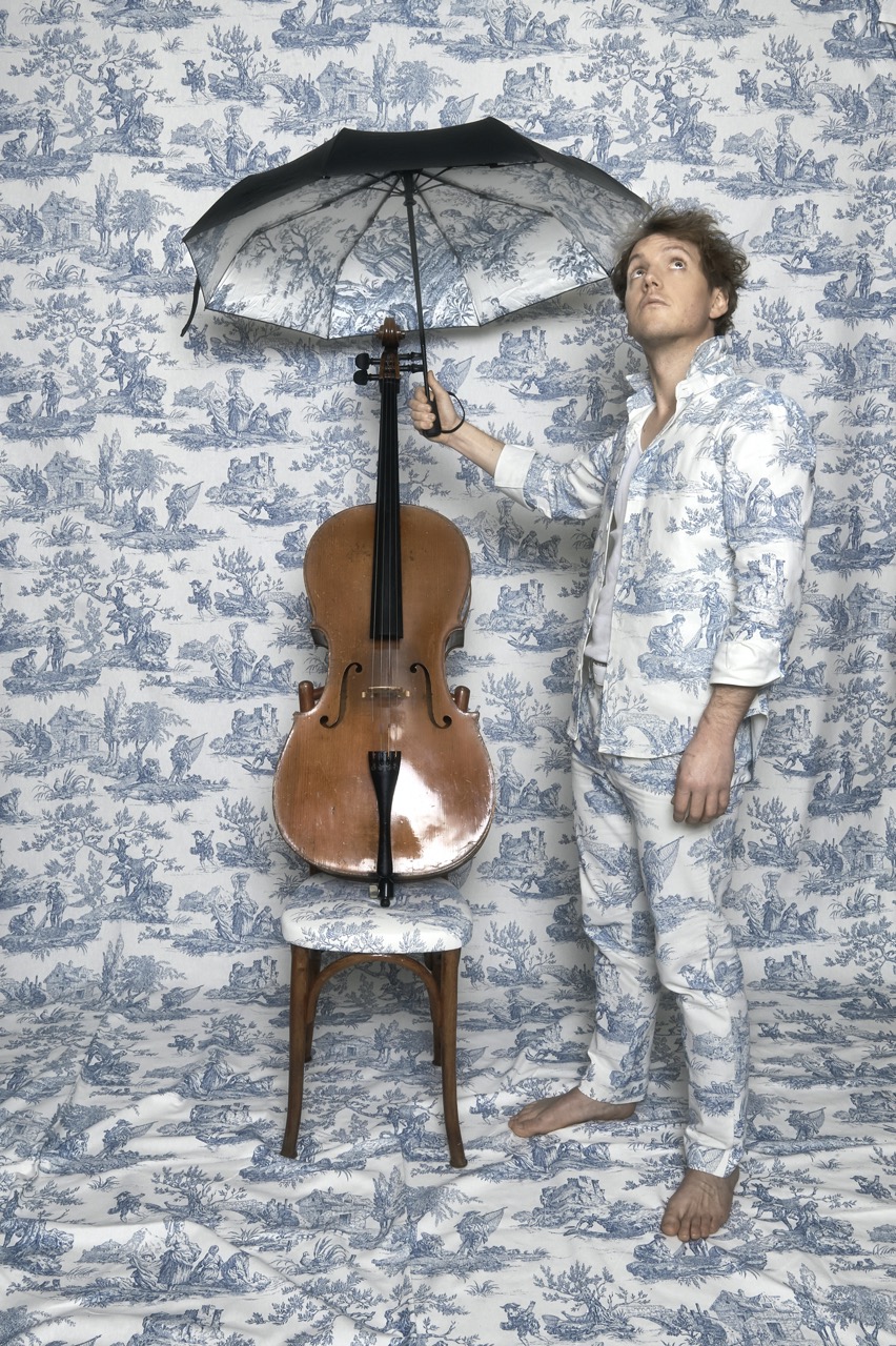 Photo d'Archipol debout tenant un parapluie à la main au dessus de son son violoncelle. Une photo d' Armelle Yons.